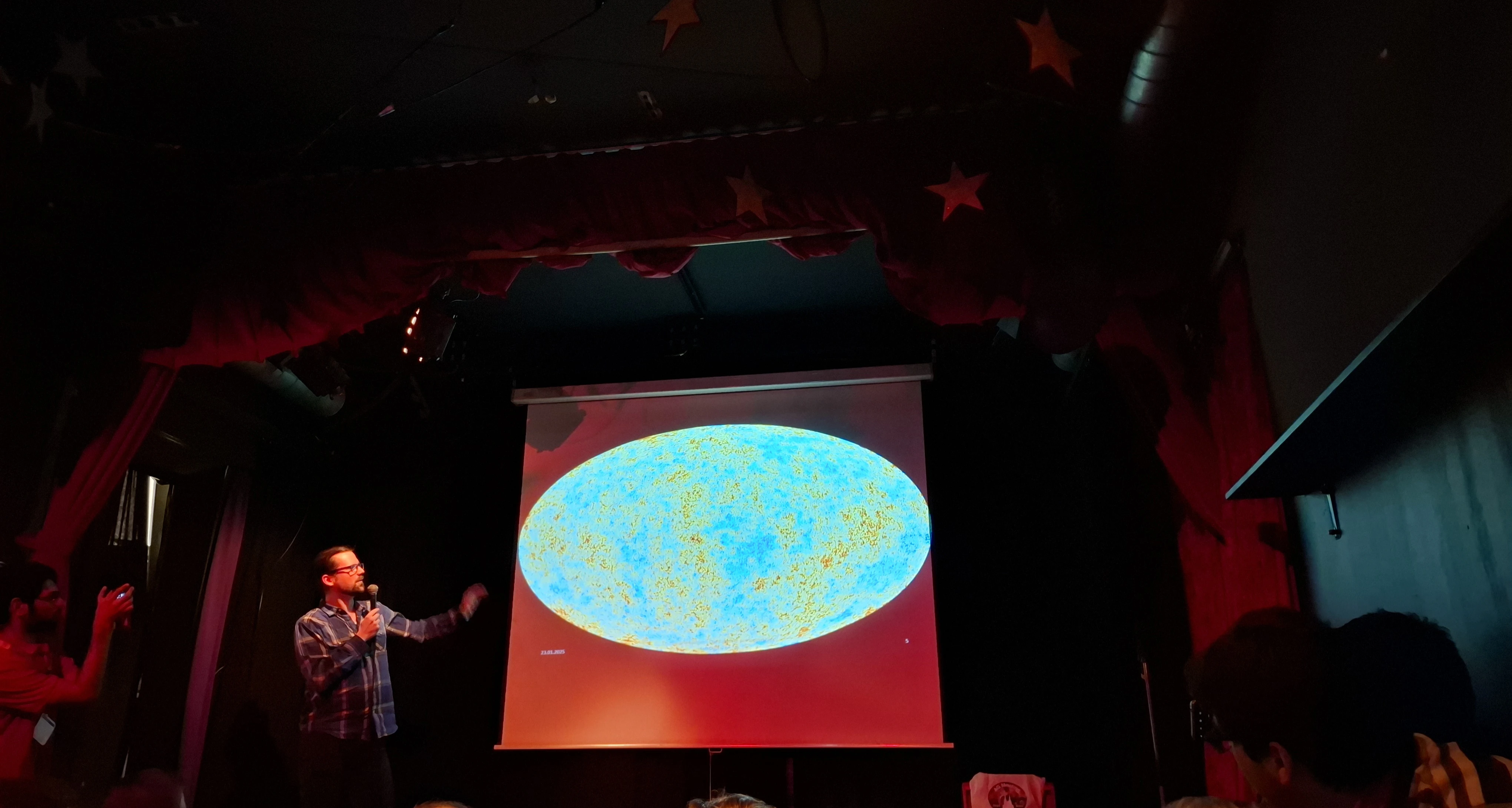 A speaker pointing to a projected all-sky map of the cosmic microwave background in a dimly lit venue.